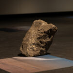 A photographic print on silk rests flat on the gallery ground with a basalt rock on top of it
