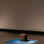 A photographic print on silk rests flat on the gallery ground with a basalt rock on top of it and a blue string hanging from the ceiling above