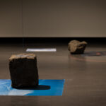 An art installation with rocks and photographic prints on silk resting on the floor and coloured strings hanging from the ceiling